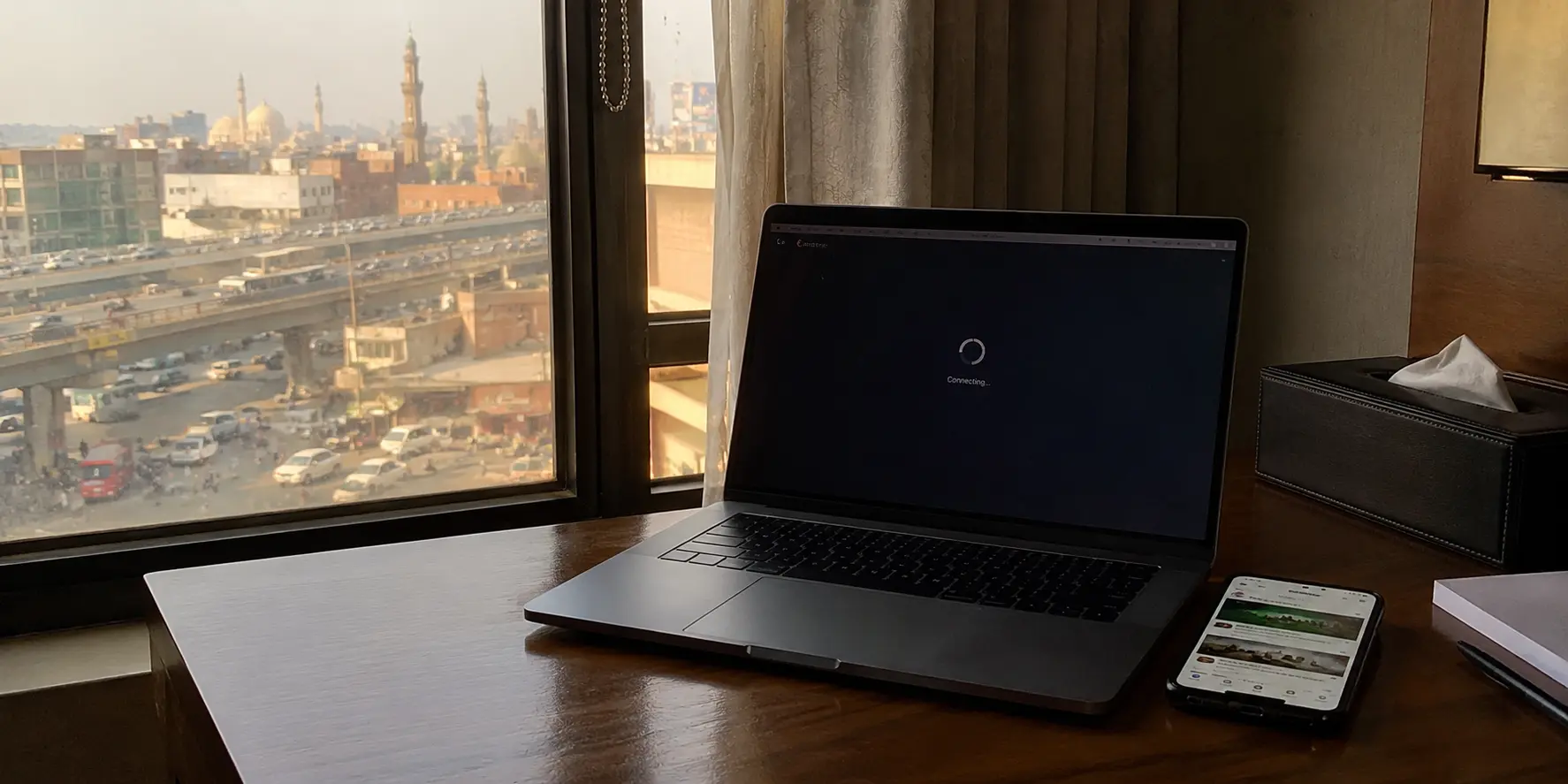 A laptop stuck connecting beside a phone in a Lahore hotel workspace, with Pakistan traffic and mosques visible outside the window