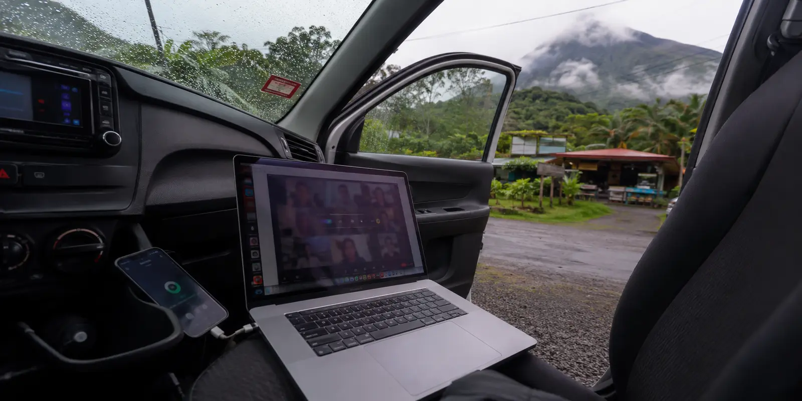 Portátil en una videollamada y teléfono compartiendo datos dentro de un automóvil detenido junto a una carretera lluviosa de Costa Rica