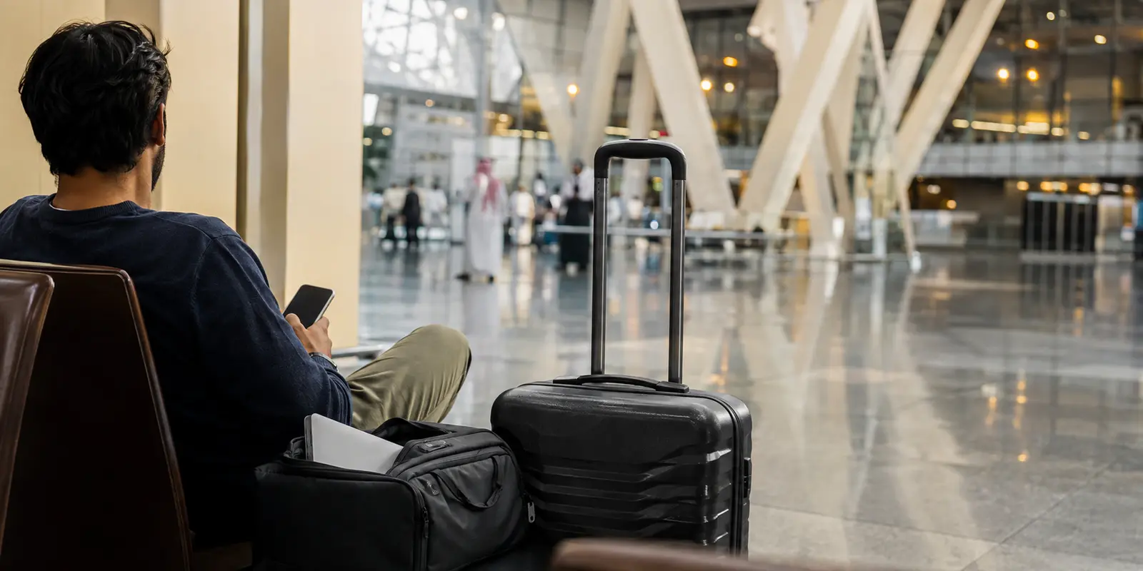 مسافر ينتظر في مطار الرياض وبجواره حقيبة سفر وهاتف وحاسوب محمول