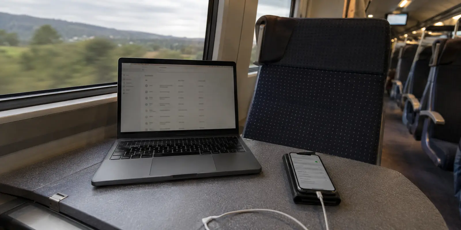 Notebook e celular conectados em uma mesa de trem, com a paisagem em movimento pela janela.