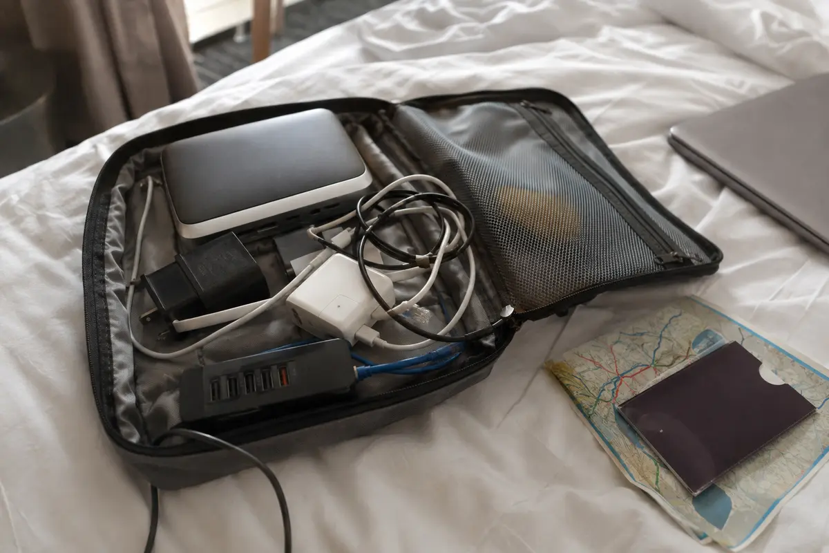 Bolsa abierta con router de viaje, adaptadores y cables enredados sobre una cama de hotel