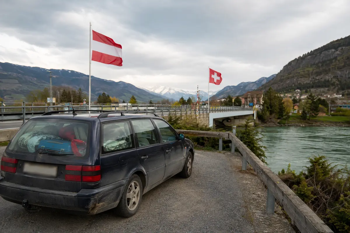 Vollgepacktes Auto zwischen österreichischer und Schweizer Flagge an der Grenze