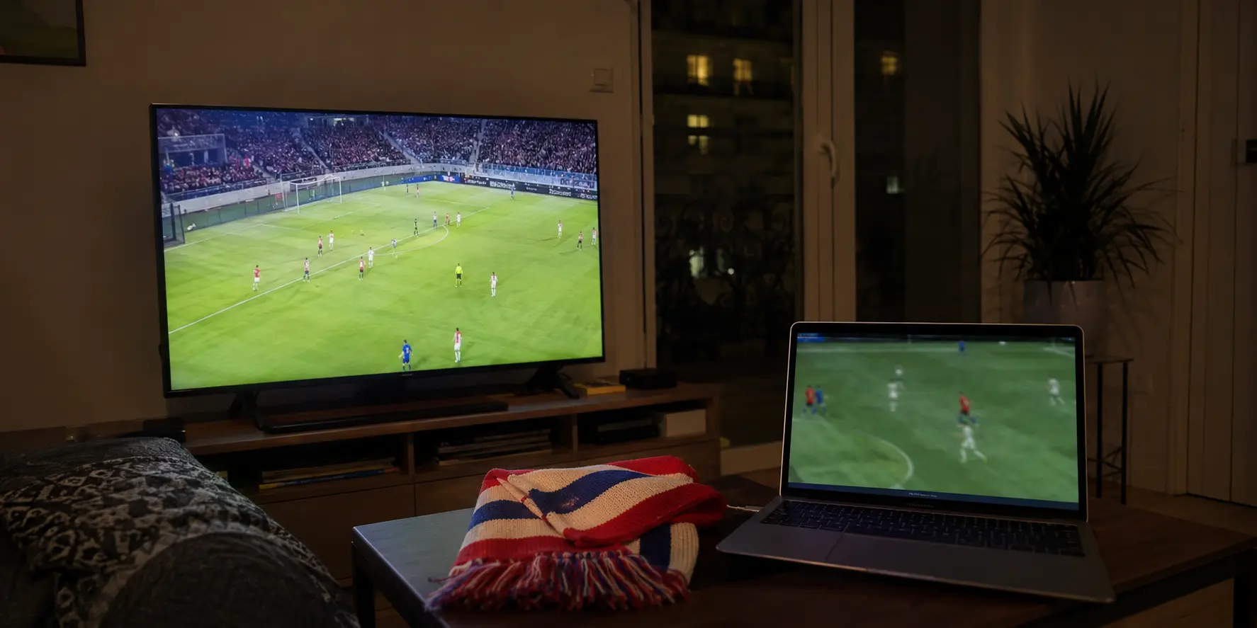 Partido de fútbol en un televisor y portátil con audio desincronizado junto a una bufanda chilena