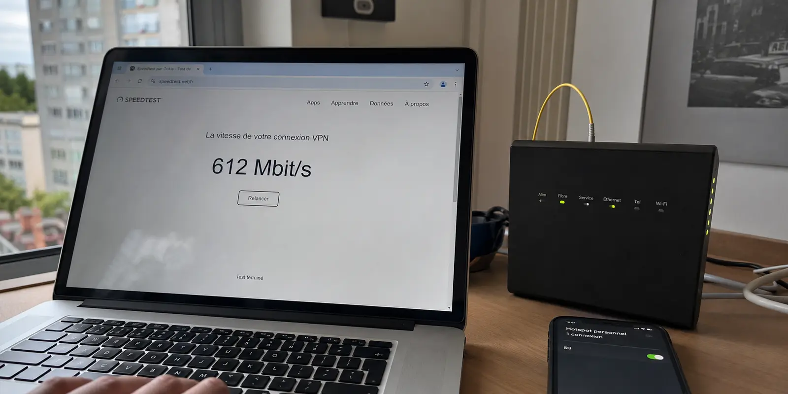 Un test affiche 612 Mbit/s sous VPN à côté de la box fibre et d’un téléphone connecté en 5G