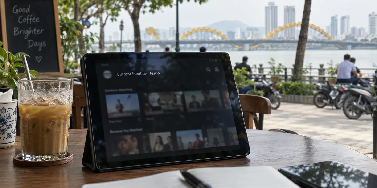 A streaming app reports Hanoi on a tablet at a riverside café in Da Nang