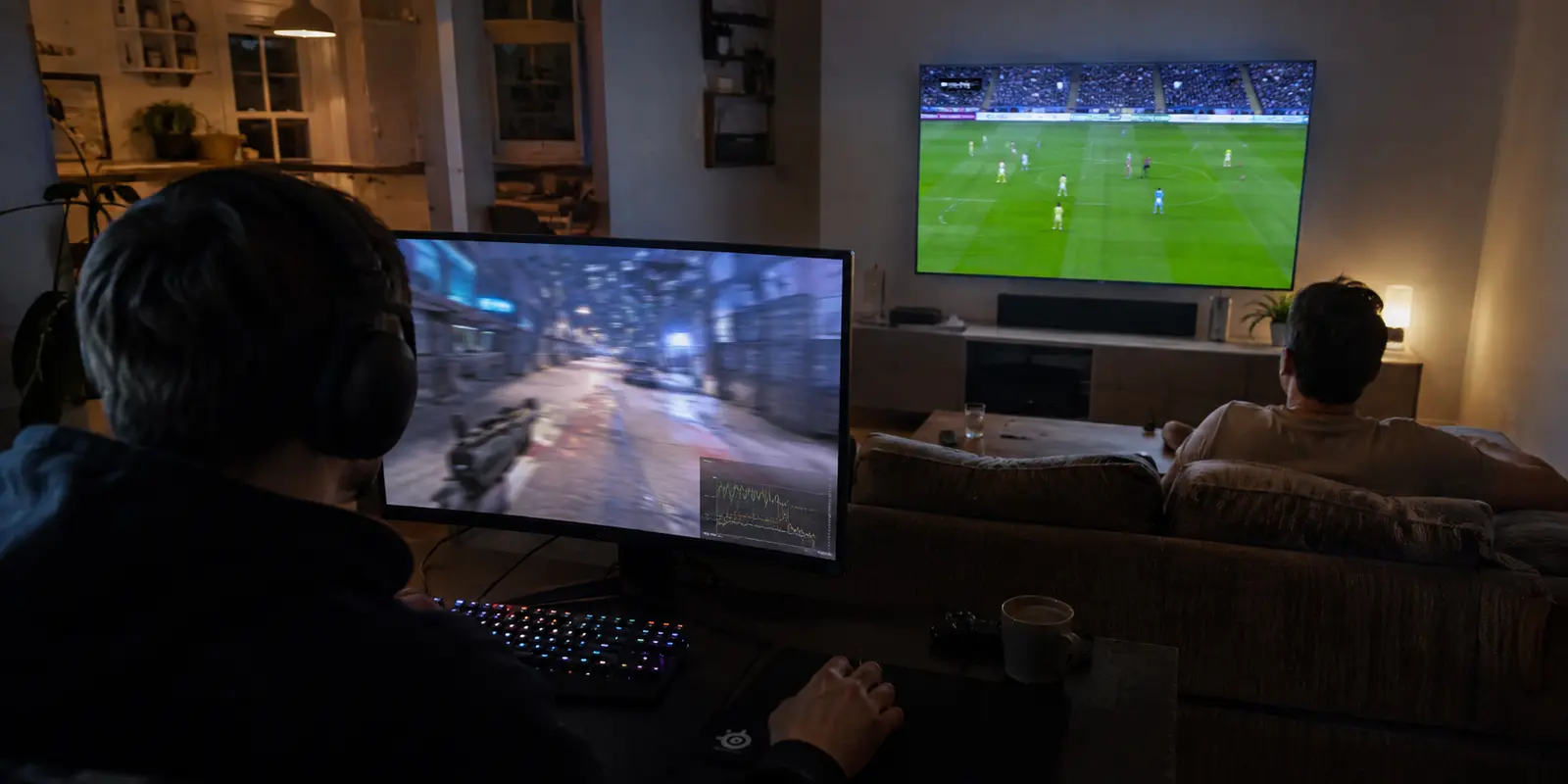 Uma pessoa joga no computador enquanto outra assiste a futebol na televisão da mesma casa