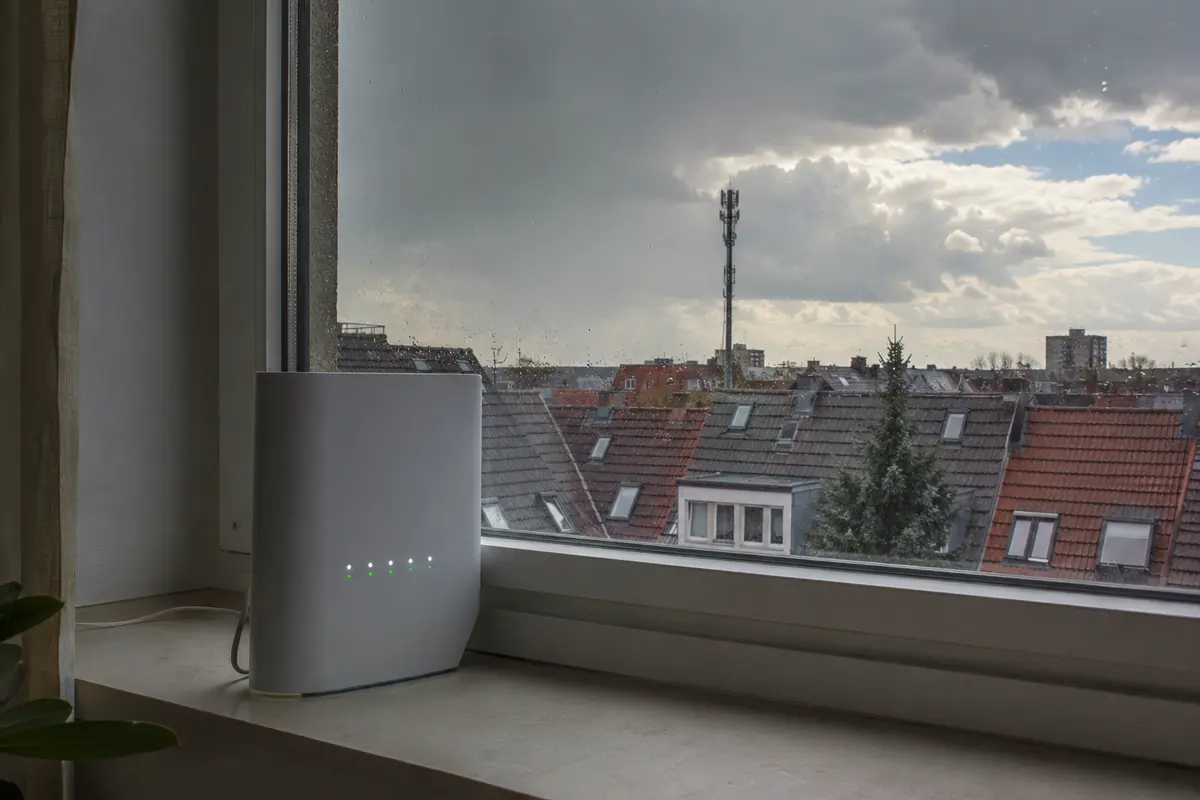 5G-HomeSpot am Fenster mit Mobilfunkmast hinter regennassen Dächern